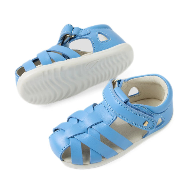 Two Baltic Sea Blue toddler leather Bobux Step Up SU Tropicana II Closed Toe Sandals with white soles. One sandal is positioned upright, revealing the top and side view, while the other is flipped to showcase the sole. The sandals have a woven design and a hook-and-loop strap, embodying classic closed-toe sandal style for extra protection and comfort., 9420074809957, 9420074809964, 9420074809971