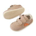 A pair of Bobux Step Up SU Champ Sneaker First Walker shoes in Warm Taupe, featuring light brown uppers with hook and loop straps, beige soles, suede toe caps, and an orange side detail. One shoe is upright while the other displays the sole., 9420074861894, 9420074861900, 9420074861917