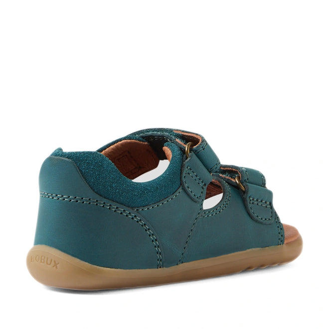 A Mediterranean green Bobux SU Step Up Driftwood First Walker Open Toe Sandal for children, designed as ideal first walking shoes, featuring leather construction with cut-outs, a tan rubber sole, and a brown interior lining. The sandal is shown from the back left angle and includes adjustable hook-and-loop fastener straps for a secure fit., 9420074807458, 9420074807465, 9420074807472
