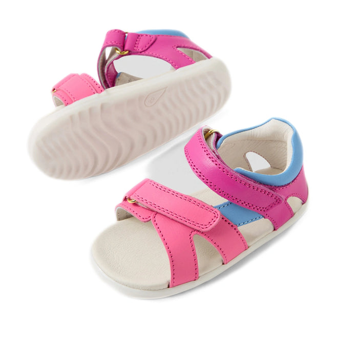 A pair of Bobux SU Step Up Dune First Walker Open Toe Sandals in Rose Bright Pink, featuring pink adjustable straps, blue accents, and a white sole. One sandal is upright while the other is shown from below to highlight the wavy texture of the sole. These charming leather sandals combine comfort and playful design for little feet, ideal for first walkers., 9420074856128, 9420074856135, 9420074856142