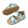 Bobux Step Up Flora First Walker Jelly Mint Metallic Blue sandal for children features open-toe metallic blue straps with moon and star embellishments, an adjustable Velcro closure, soft tan cushioned insole, and a light brown rubber sole. One leather sandal is displayed upright while the other showcases the sole.