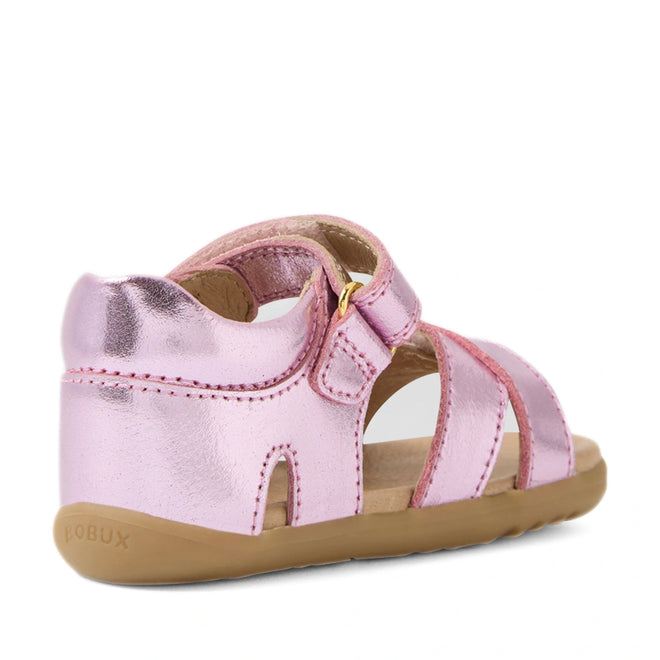 Introducing the Bobux Step Up Flora First Walker Sandal in Pink Lemonade Metallic—a stylish metallic pink toddler sandal featuring a closed heel, open toe, and adjustable strap. Perfect for little first walkers, these smooth leather sandals have a light brown sole with visible stitching and are displayed at an angle to highlight both the back and side design.