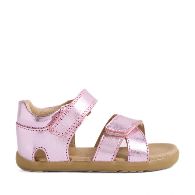The Bobux Step Up Flora First Walker Sandal in Pink Lemonade Metallic is a children’s metallic pink leather sandal, featuring a closed heel, two adjustable Velcro straps, an open toe, and a tan rubber sole. It is displayed in a left side profile on a white background.
