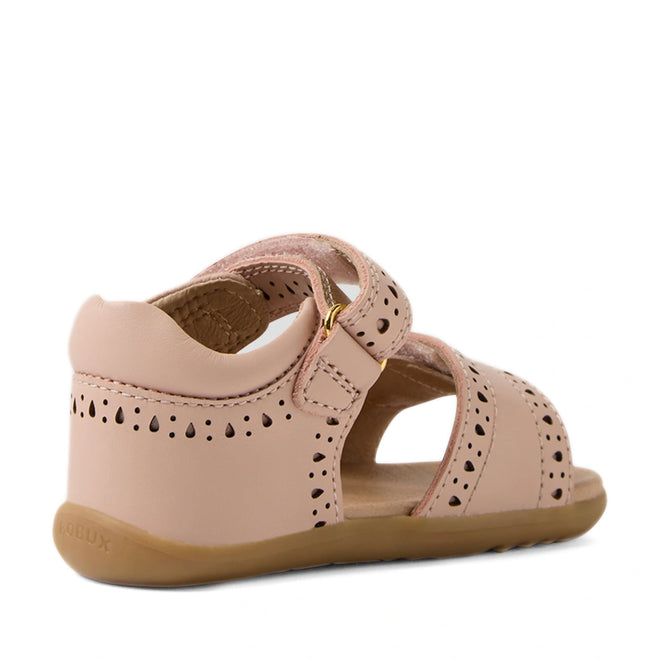 Bobux Step Up Grass Logan First Walker Sandal in Cameo Rose Pink—first walker girls’ leather sandals with decorative cutouts, a closed heel, open toe, ankle strap with gold buckle, and a flexible tan rubber sole; shown from the back left side.