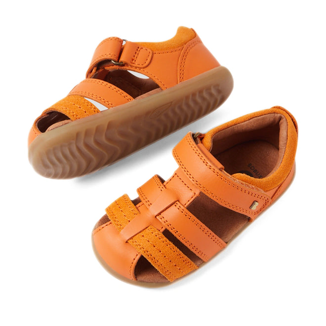 The Bobux Step Up Sandal Roam Closed Toe Sandals in Orange Peel are displayed on a white background. One sandal stands upright while the other is tilted to reveal the tread pattern, showcasing the quality and design of these toddler leather sandals with Velcro straps and rubber soles.