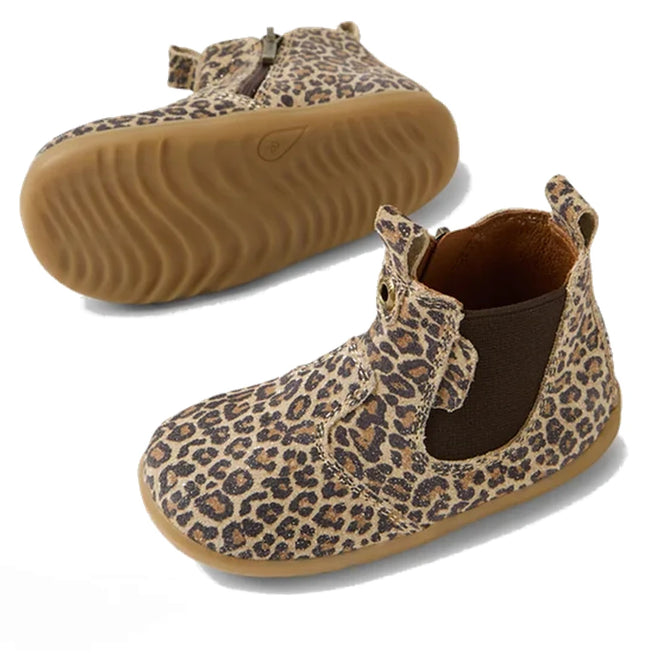 The Bobux Step Up Jodhpur Boot Leopard Shimmer features a brown rubber sole, leopard print suede/nubuck leather, a side zipper, and wavy tread—one kid's heritage boot stands upright while the other displays its sole.