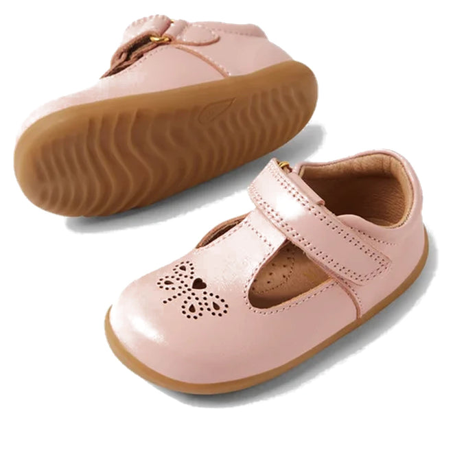The Bobux Step Up T-Bar Rosie First Walker Shoe in Ballet Pink features pink leather, tan rubber soles, a T-strap, and heart and flower cutouts on the toe. One Mary Jane flat is upright while the other displays its sole., 51161, 9420074849946, 9420074849953, 9420074849960