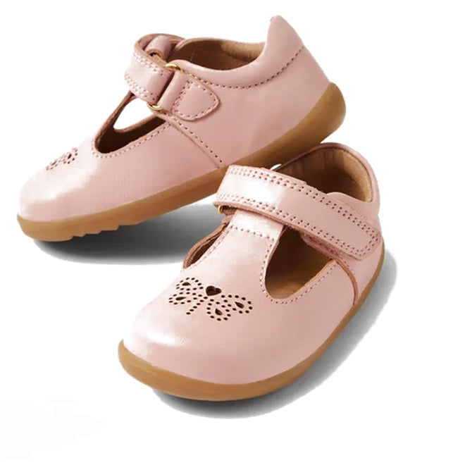 The Bobux Step Up T-Bar Rosie First Walker Shoe in Ballet Pink features light pink leather, tan soles, a bow cutout on the toe, and T-bar straps—perfect toddler first walker shoes shown on a white background.