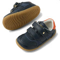 The Bobux SU Step Up Hunter Leather Toddlers Sneaker in navy features brown rubber soles, double Velcro straps, perforated toes, and orange heel accents. One shoe is upright; the other is shown from the side., 9420074833853, 9420074833860, 9420074833877