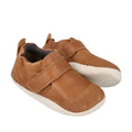 The Bobux Explorer Marvel Caramel Dress Shoe First Walker is a brown leather baby shoe with white rubber soles, hook-and-loop straps, rounded toe, soft flexible material, and scuff-resistant design for active toddlers.