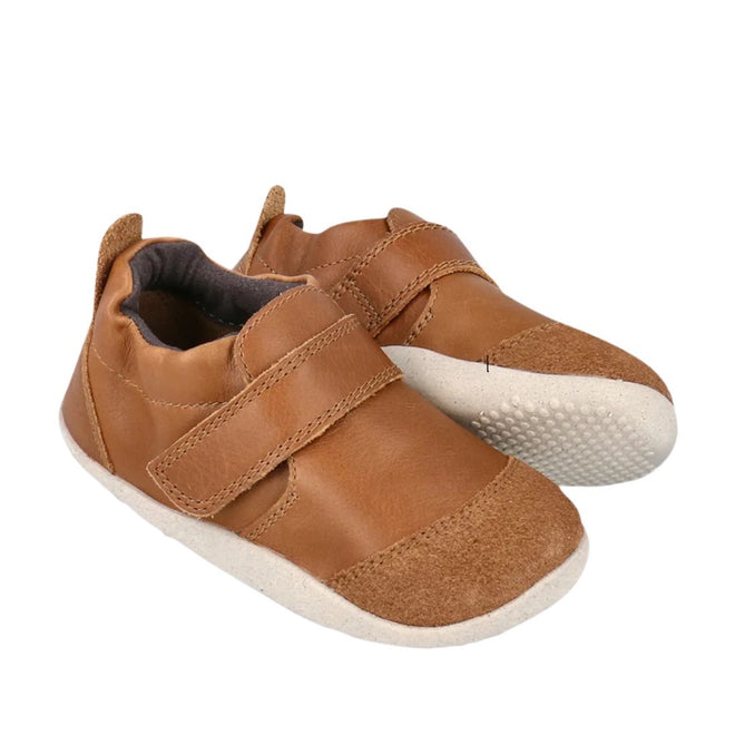 The Bobux Explorer Marvel Caramel Dress Shoe First Walker is a brown leather baby shoe with white rubber soles, hook-and-loop straps, rounded toe, soft flexible material, and scuff-resistant design for active toddlers.