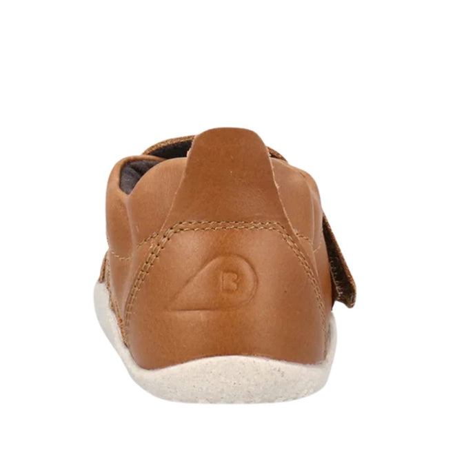 A back view of the Bobux Explorer Marvel Caramel Dress Shoe First Walker shows its brown leather, white sole, stitched logo on the heel, and side strap.