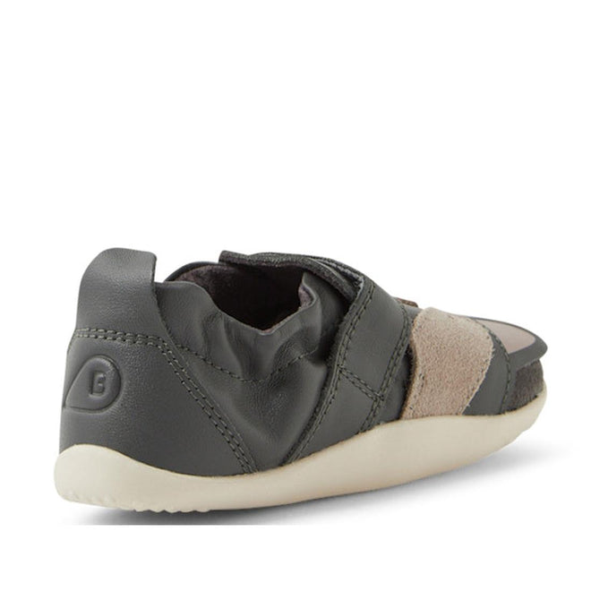 The Bobux XP Xplorer Scrunch First Walking Shoes in Smoke Grey are pre-walker toddler shoes in dark gray with beige suede accents, shown from a rear-side angle. They feature an adjustable strap, flexible material, and a light-colored textured sole., 9420074805850, 9420074805867, 9420074805874, 9420074805881, 9420074805843, 9420074805850, 9420074805867, 9420074805874, 9420074805881