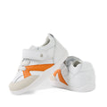 Two Bobux Xplorer XP Chase White Orange Peel First Walker Crawler shoes are shown—white toddler sneakers featuring orange accents, velcro straps, and beige toe patches. Designed for pre-walkers with a flexible sole, they provide comfort and support for little feet. One shoe stands upright while the other is positioned on its side to reveal the detailed sole and side features.