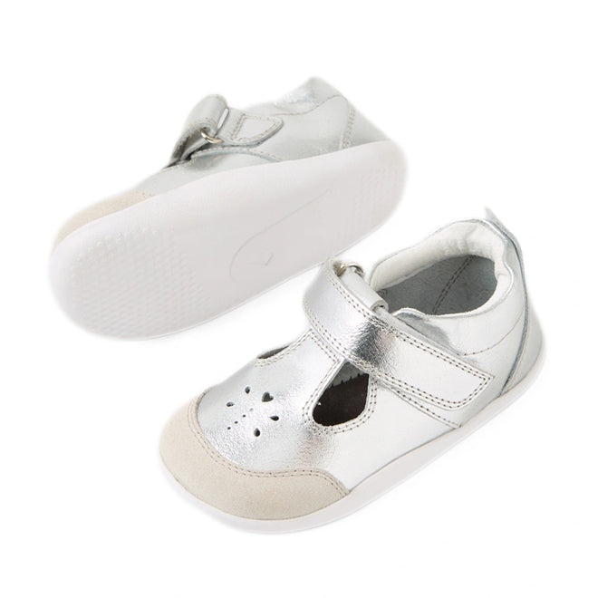 Bobux XP Explorer Rue First Walker Sandal Silver—this pair of shiny silver toddler shoes features white soles, beige toe caps, cutout detailing on the front, and easy hook-and-loop (Velcro) straps. Perfect as first walking or pre-walker shoes; one shoe is upright while the other lies on its side to display the sole.