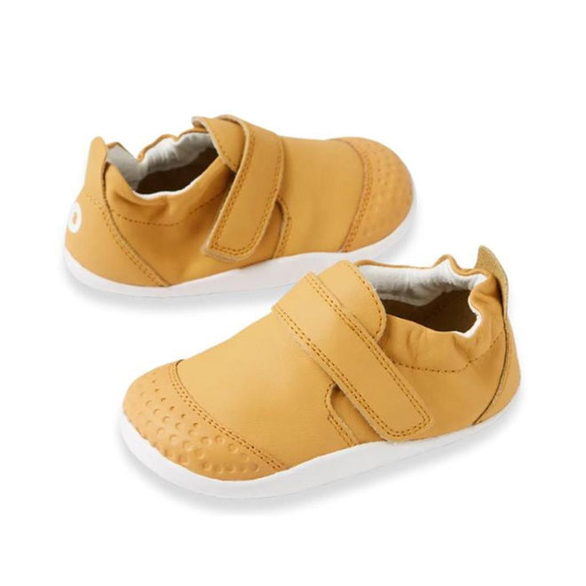 The Bobux XP Xplorer First Walking Shoes Go Oak Buff Tan are small tan pre-walker leather shoes with white flexible soles, velcro straps, and textured toe caps—perfect for your baby's first steps and shown against a white background.