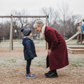 An older woman in a red coat smiles warmly at a young boy wearing a blue jacket, hat, and Bobux IW Iwalk Breakpoint Leather Coronet Blue sneakers as they face each other in a playground on a chilly day with swings in the background., 9420074817709, 9420074817716, 9420074817723, 9420074817730