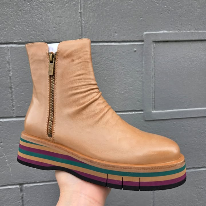 The Django & Juliette Ganni Cognac Tan Green Leather Heeled Boot features a gold zipper and thick platform sole with black, green, purple, and brown layered stripes, shown against a gray textured wall.
