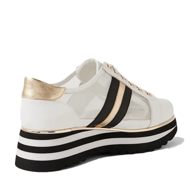 The Django & Juliette Eminta Platform Shoe is a white women’s sneaker featuring sheer leather side panels, black and gold stripes, a gold heel accent, and a thick black-and-white striped platform sole.