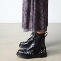A person wearing a long plaid skirt and shiny black Dr Martens 1460 Hardware HDW Buttero Leather Boots stands on a light-colored floor., 49862, 190665797572, 190665797589, 190665797602, 49866, 49867, 49868, 49869