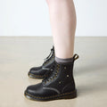 A person wearing black Dr. Martens Women's 1460 Pascal Flower Black Grizzly Leather Boots with floral details and gray socks stands on a light-colored floor against a plain white wall., 52216, 190665781137, 190665781144, 190665781151, 190665781175, 190665781182, 52222