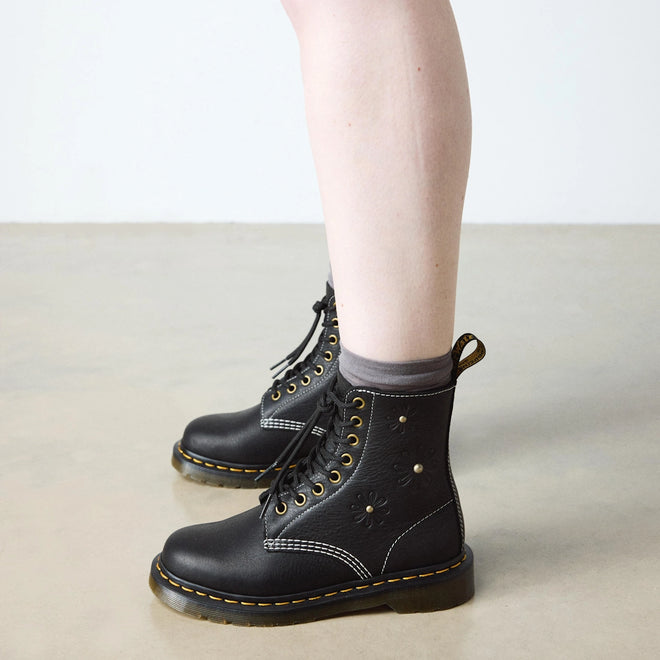 A person wearing black Dr. Martens Women's 1460 Pascal Flower Black Grizzly Leather Boots with floral details and gray socks stands on a light-colored floor against a plain white wall., 52216, 190665781137, 190665781144, 190665781151, 190665781175, 190665781182, 52222