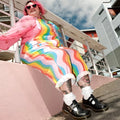 A person with pink hair and heart-shaped sunglasses sits on a pink ledge, wearing a rainbow-striped jumpsuit, lace socks, and Dr Martens 8065 2 Strap Mary Jane Shoe Black Smooth, smiling with modern buildings in the background., 883985051768, 883985051775, 883985051782, 883985051799, 883985051805, 883985051812
