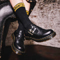 Close-up of a person wearing Dr Martens 8065 2 Strap Mary Jane Shoe Black Smooth with yellow stitching and black socks with yellow text, seated on a black leather chair against a white brick wall., 883985051768, 883985051775, 883985051782, 883985051799, 883985051805, 883985051812