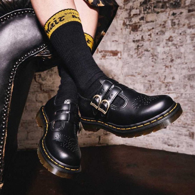 Close-up of a person wearing Dr Martens 8065 2 Strap Mary Jane Shoe Black Smooth with yellow stitching and black socks with yellow text, seated on a black leather chair against a white brick wall., 883985051768, 883985051775, 883985051782, 883985051799, 883985051805, 883985051812