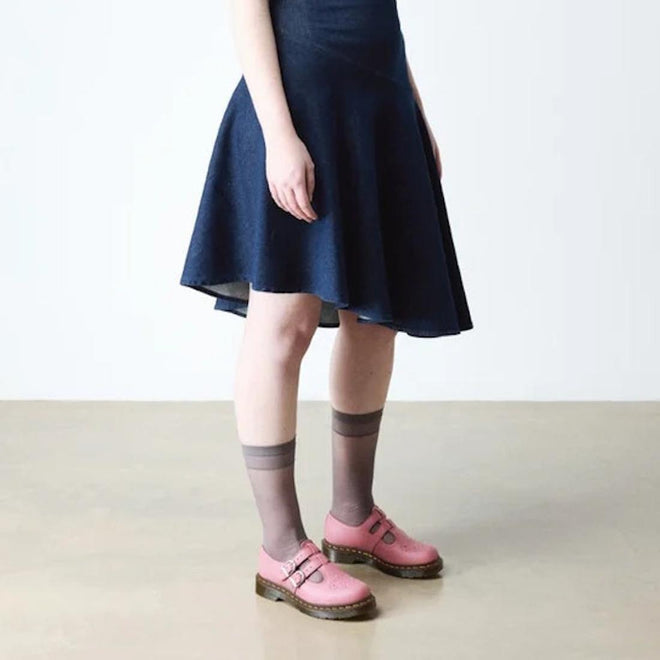 A person wearing a dark blue dress, sheer gray ankle socks, and Dr. Martens 8065 2 Strap Mary Jane Shoe in Dusky Rose Pink stands on a beige floor against a plain white background., 50241, 50242, 50243, 50244, 50245, 50246, 50247, 50241, 190665781861, 190665781878, 190665781892, 50245, 50246, 50247