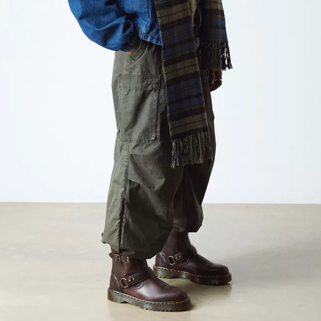 A person wearing Dr Martens Anistone Harness Bex Crazy Dark Brown Horse Biker Boots, olive cargo pants, a blue jacket, and a plaid fringe scarf stands on a light-colored floor against a white wall., 49399, 190665745689, 190665745696, 190665745719, 49407, 49409, 49411, 49412