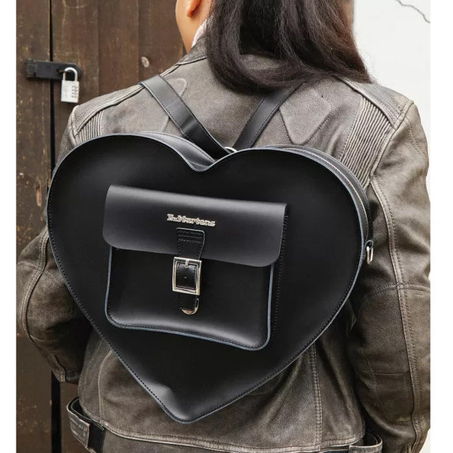 Wearing a gray leather jacket and Dr. Martens, someone carries the Dr Martens Large Big Heart Box Backpack Leather Black with a front pocket and buckle, standing by a wooden door with a small padlock., 190665665406