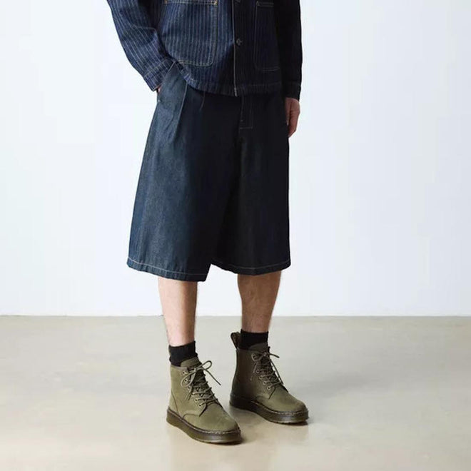 A person wearing a pinstriped jacket, dark knee-length denim shorts, black socks, and Dr. Martens Brookline Chukka Boots in Olive Milled Nubuck Leather with a lightweight outsole stands on a light-colored floor against a plain white background., 51342, 190665797053, 190665797060, 190665797084, 190665797091, 190665797107, 190665797121, 51349