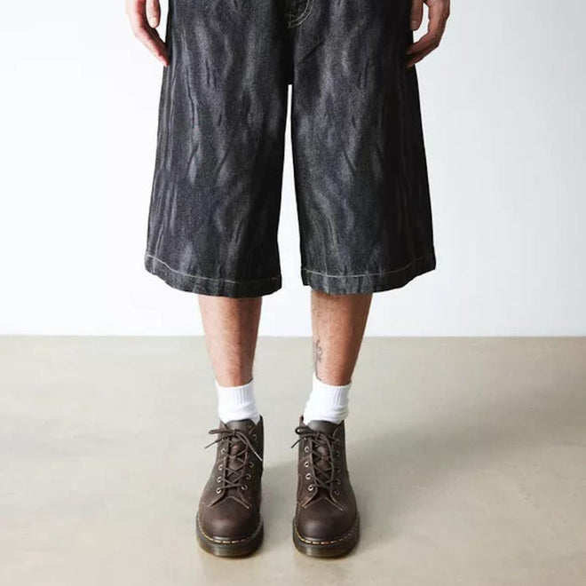 A person wearing long, loose black denim shorts, white socks, and Dr Martens Church Monkey Boot Crazy Horse Dark Brown stands on a light floor against a plain white wall. Only their legs are visible, highlighting a vintage-inspired look., 44507, 190665703580, 190665703597, 190665703610, 190665703627, 190665703634, 190665703658, 44514, 44515