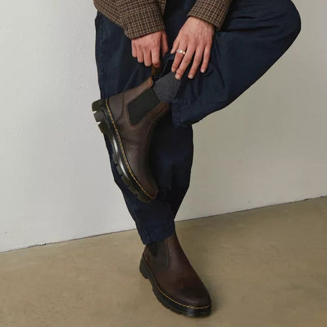 A person leaning against a wall with one leg angled against the others knees, wearing Dr Martens Embury boots in crazy horse brown showing the black elastic gussets,  and yellow stitching around the sole .,
190665358643,
190665358650,
190665358674,
190665358681,
190665358698,
190665358711,