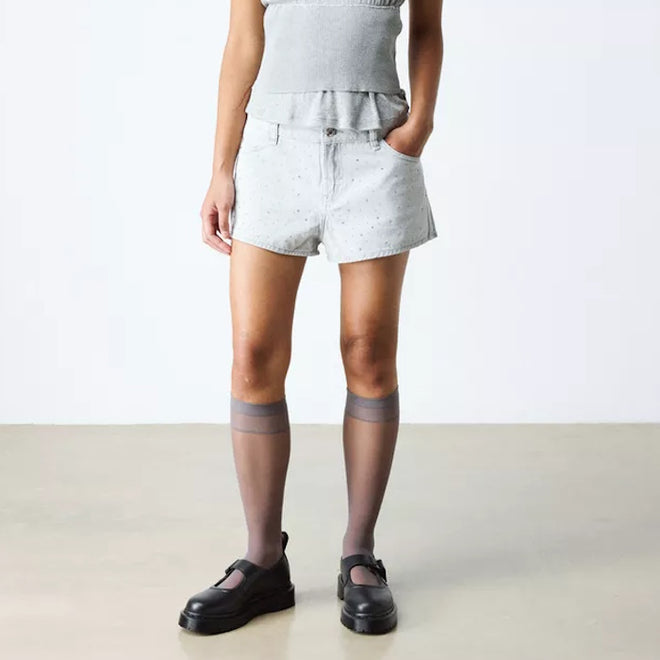 A person wears light gray shorts, a matching top, sheer knee-high socks, and Dr Martens Bex Indica II Mary Jane Tech Nappa Mono Black shoes against a plain background. With one hand in their pocket, their face remains out of view., 190665804744, 190665804751, 190665804768, 190665804782, 190665804799, 190665804805, 50377
