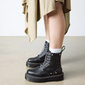 A person wearing Dr Martens Jadon WDS White Stitching Hardware 8 Eye Boot Black with thick soles, white socks, and a beige asymmetrical skirt stands on a light floor, highlighting the platform silhouette., 50347, 50348, 190665775471, 190665775488, 190665775501, 50353, 50354, 50355, 50356