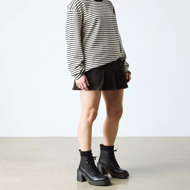 A person in a black-and-white striped long-sleeve shirt, black pleated skirt, black socks, and Dr Martens Jesy Sendal 6 Tie Flared Heel Boots stands against a plain background, with their face not visible., 190665511796, 190665511802, 190665511819, 190665511833, 190665511840, 190665511857