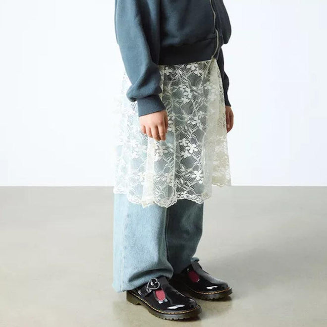 A person stands indoors on a polished floor wearing light blue jeans, a dark blue jacket, a sheer white lace skirt overlay, and Dr Martens kids - Polley II Heart T Bar Junior Black Patent shoes. The photo is cropped at the shoulders., 50432, 50433, 190665786743, 190665786750, 190665786651, 190665786675, 190665786699