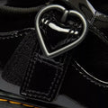 Close-up of the Dr Martens kids Polley II Heart T Bar Junior in black patent, showing a shiny strap with a heart-shaped buckle, detailed stitching, and glossy finish—perfect classic-inspired school shoes., 50432, 50433, 190665786743, 190665786750, 190665786651, 190665786675, 190665786699
