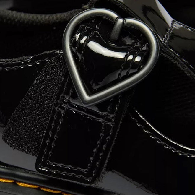 Close-up of the Dr Martens kids Polley II Heart T Bar Junior in black patent, showing a shiny strap with a heart-shaped buckle, detailed stitching, and glossy finish—perfect classic-inspired school shoes., 50432, 50433, 190665786743, 190665786750, 190665786651, 190665786675, 190665786699