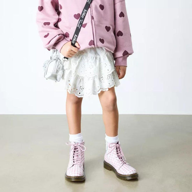 A child in a pink heart-print sweatshirt, white eyelet skirt, white socks, and Dr Martens kids - 1460 J Juniors Gingham Pink Lace Up Boots with zip stands on a light floor with an Athena leather crossbody bag against a plain background., 50424, 50425, 50426, 190665779165, 190665779066, 190665779080, 190665779103