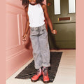 A child stands on a patterned rug by a green door, wearing a white tank top, gray cuffed jeans, and Dr Martens Kids 1460 Junior Pink Glitter Ombre Gradient boots, with one arm slightly bent and their face partially out of frame.