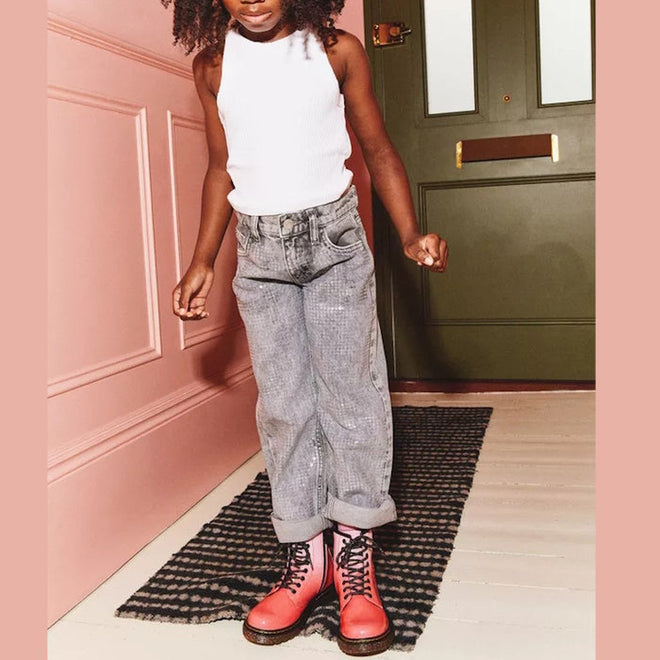 A child stands on a patterned rug by a green door, wearing a white tank top, gray cuffed jeans, and Dr Martens Kids 1460 Junior Pink Glitter Ombre Gradient boots, with one arm slightly bent and their face partially out of frame.