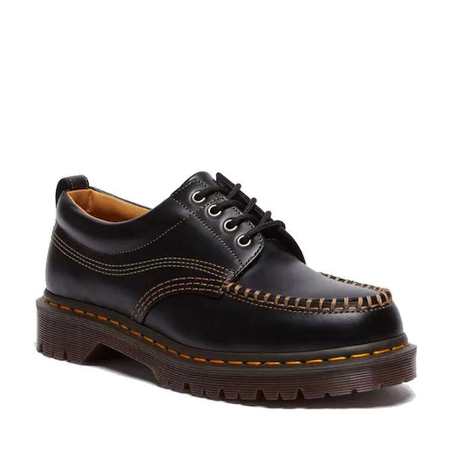 The Dr Martens Lowell Moc Toe Black Leather Shoe features thick brown stitching, a chunky sole, Goodyear welt construction, and yellow edge detailing, shown from an angle highlighting the side and front.