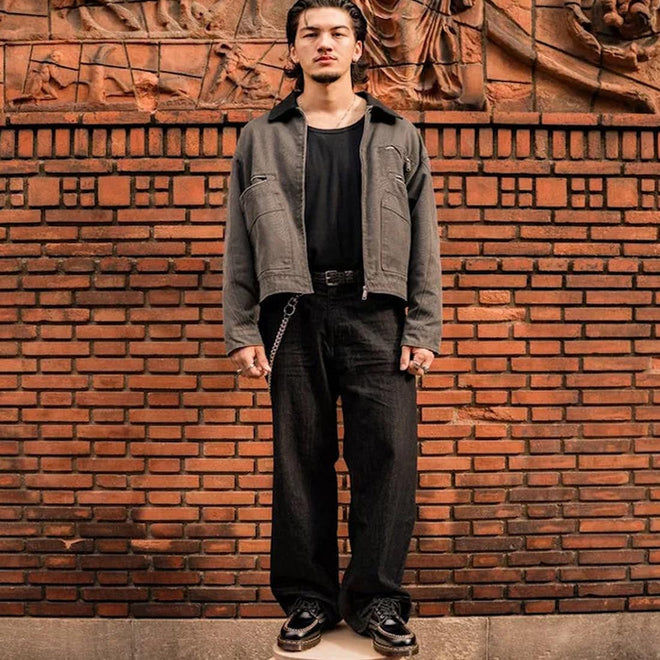 A man stands before a decorative brick wall, wearing a black t-shirt, gray jacket, loose black pants, and Dr Martens Lowell Moc Toe Black Leather Shoes with a chain detail. His hands are at his sides as he looks straight at the camera., 50395, 50396, 50397, 50398, 50399, 190665668940, 190665668957, 190665668971, 190665668988, 50404