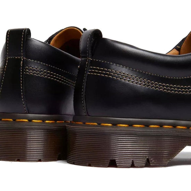 Close-up of the back and side of two Dr Martens Lowell Moc Toe Black Leather Shoes in aniline leather, featuring thick brown ridged soles and signature yellow stitching., 50395, 50396, 50397, 50398, 50399, 190665668940, 190665668957, 190665668971, 190665668988, 50404
