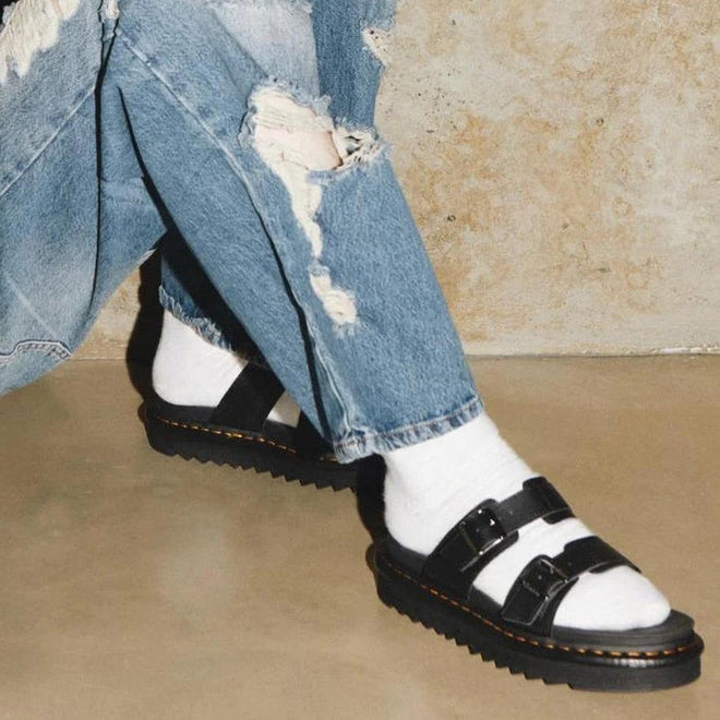 A person sitting on the ground, only the legs and feet in the shot, wearing blue denim jeans , white socks and Dr Martens Myles black leather slide sandals with 2 straps across the foot with buckle on the outer foot for adjustment.,
190665653779,
190665653786,
190665653793,
190665653816,
190665653823,
190665653830,