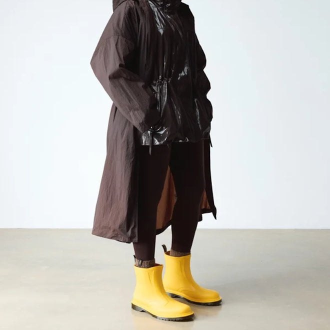 A person wears a long dark brown raincoat with a shiny upper, dark pants, and bright yellow Dr Martens 1460 Sub Max Pull On Rain Boot Yellow PVC, standing indoors against a plain light background., 49435, 190665746464, 190665746471, 190665746495, 49440, 49441, 49442