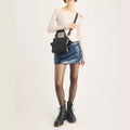 A person wearing a fitted white top, blue mini skirt, sheer tights, and black lace-up boots crafted from sleek black Kiev leather stands against a plain light background, carrying the Dr Martens Small Keiv Leather Tote Bag Black over their shoulder.